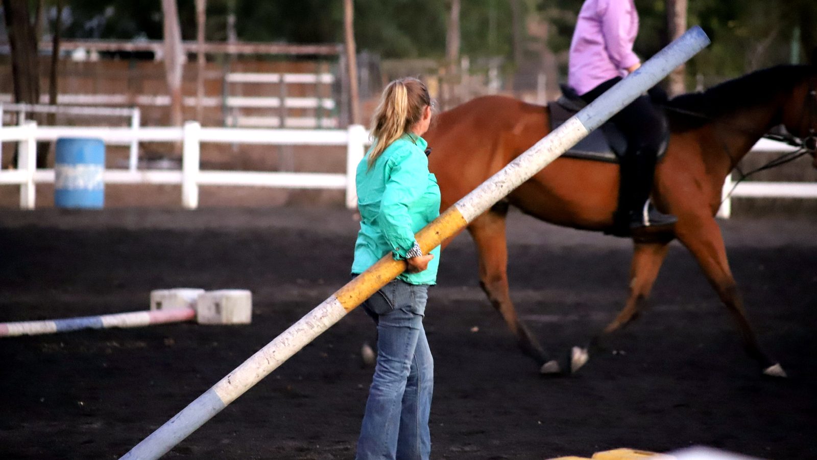A coach carrying a painted ground pole across an arena, a rider working a bay horse behind her