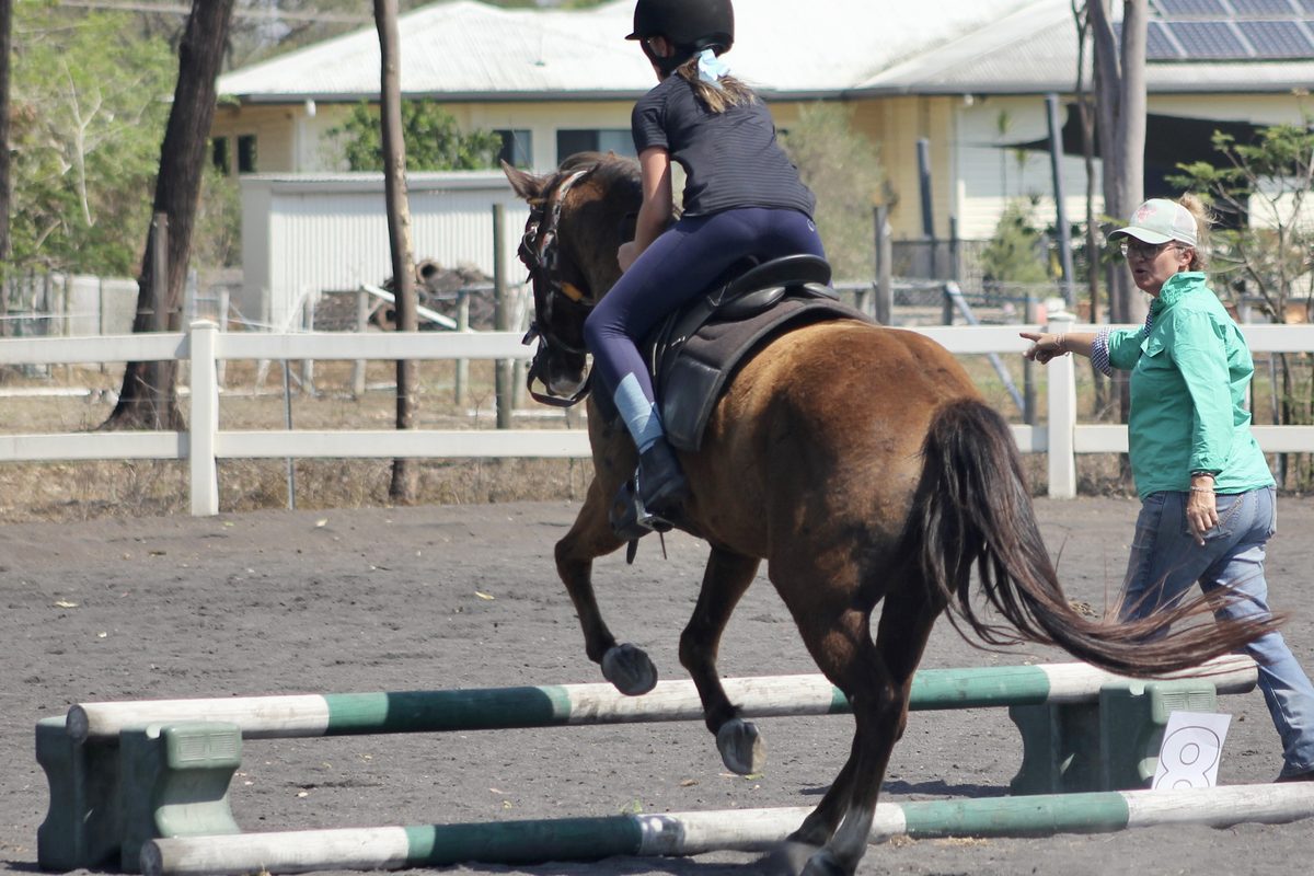 Tania giving a correction, arm outstretched, as a rider takes her horse over a low jump